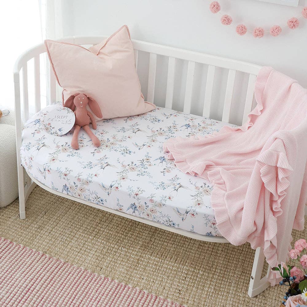 White crib with floral bedding, pink pillow, and blanket in a nursery setting.