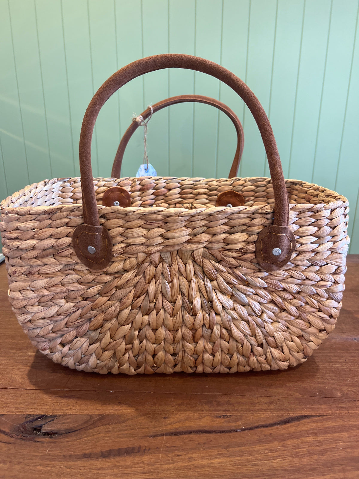 Woven handbag with brown handles on a wooden surface against a light green wall.
